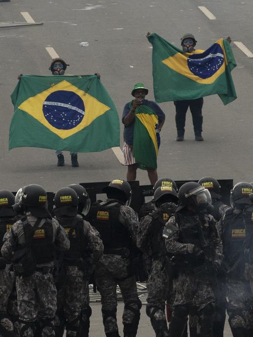 Anhänger des Ex-Präsidenten Bolsonaro stehen, brasilianische Fahnen haltend, Sicherheitskräften gegenüber. 