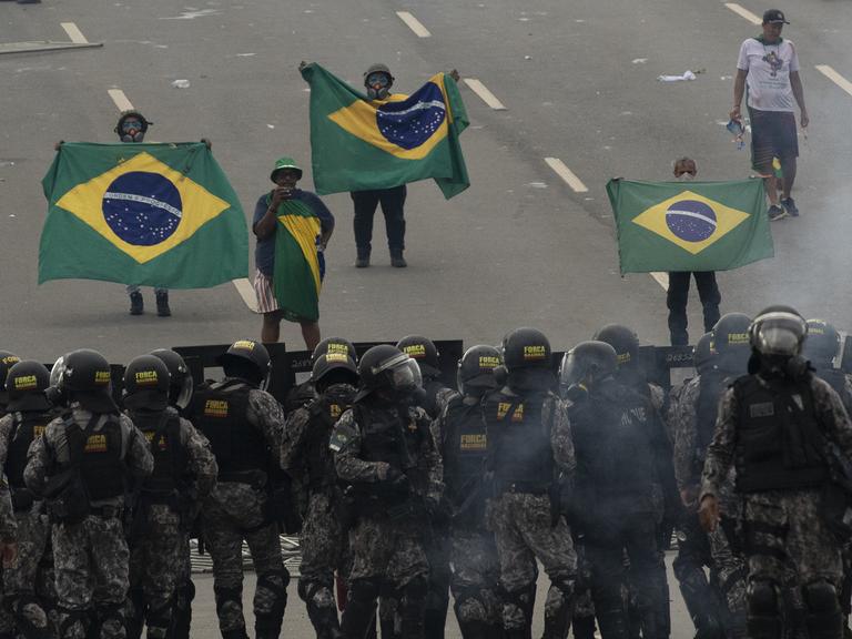 Anhänger des Ex-Präsidenten Bolsonaro stehen, brasilianische Fahnen haltend, Sicherheitskräften gegenüber. 