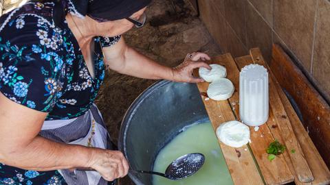 Halloumi Käseherstellung in einem traditionelles Haus im kleinen Dorf Letymbou in Zypern. Ein Frau in Küchenschürze und Kopftuch greift nach einem weissen Käsestück, in der anderen Hand hält sie eine Kelle über einem großen Topf.
