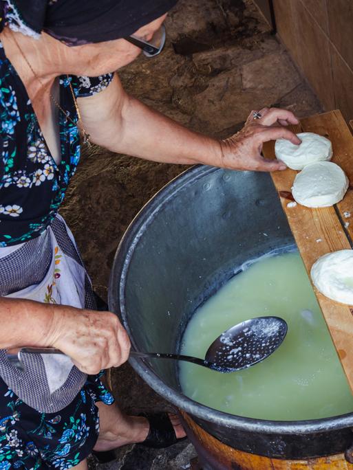 Halloumi Käseherstellung in einem traditionelles Haus im kleinen Dorf Letymbou in Zypern. Ein Frau in Küchenschürze und Kopftuch greift nach einem weissen Käsestück, in der anderen Hand hält sie eine Kelle über einem großen Topf.