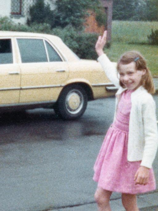 Farbige historische Filmaufnahme eines Mädchens im rosa Kleid was einem Mann in einem gelben Mercedes winkt.