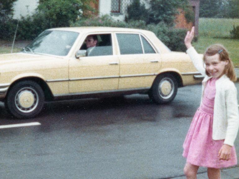 Farbige historische Filmaufnahme eines Mädchens im rosa Kleid was einem Mann in einem gelben Mercedes winkt.