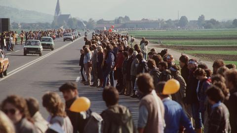 Am Rand einer Autobahn fassen sich zahlreiche Menschen an den Händen und bilden aus Protest eine Menschenkette.