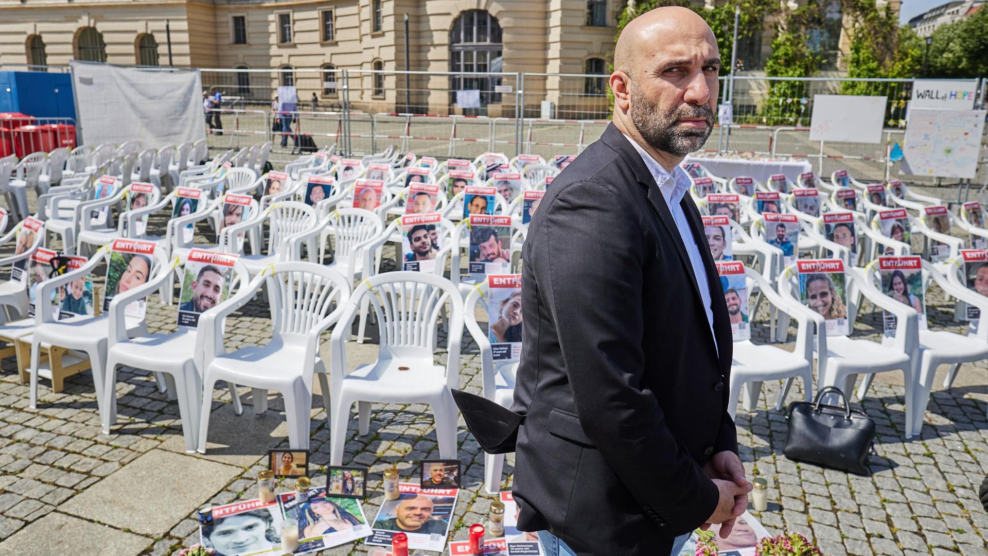 Der Psychologe Ahmad Mansour  steht vor leeren Stühlen auf denen Fotos von Hamas-Geiseln liegen. 