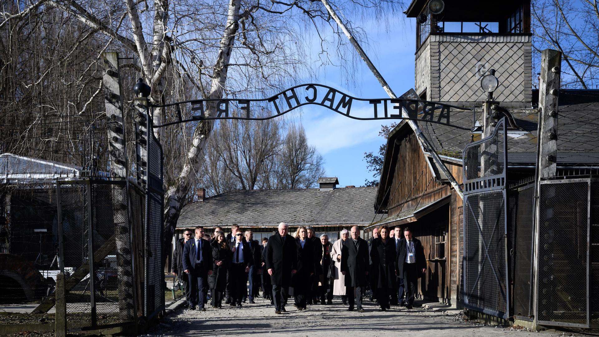 Eine Gruppe von dunkel gekleideten Menschen läuft durch das Tor in Auschwitz. Man kann den gespiegelten Schriftzug "Arbeit macht frei" über dem Tor lesen