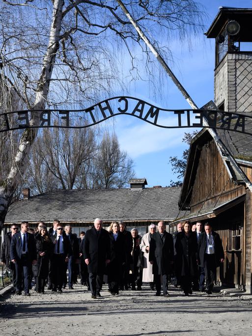 Eine Gruppe von dunkel gekleideten Menschen läuft durch das Tor in Auschwitz. Man kann den gespiegelten Schriftzug "Arbeit macht frei" über dem Tor lesen