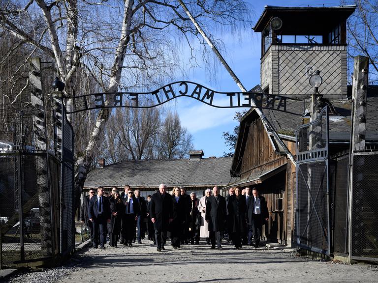 Eine Gruppe von dunkel gekleideten Menschen läuft durch das Tor in Auschwitz. Man kann den gespiegelten Schriftzug "Arbeit macht frei" über dem Tor lesen