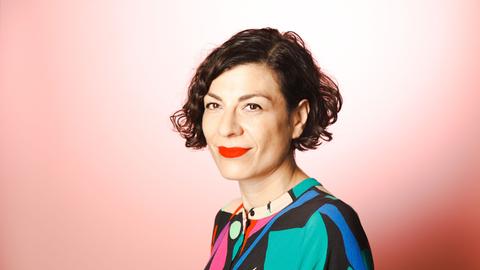 Zu sehen ist Elena Kountidou, Geschäftsführerin der Neuen deutschen Medienmacher*innen. Sie trägt kurzes, braunes welliges Haar, roten Lippenstift und ein buntes Oberteil vor einem rötlichen Hintergrund.