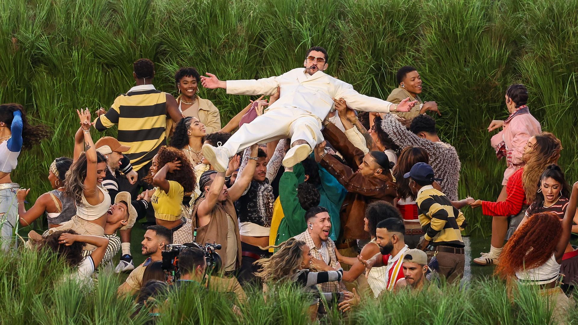 Eine Gruppe von Tänzerinnen und Tänzern steht vor Zuckerrohrpflanzen und trägt Bad Bunny über sich 