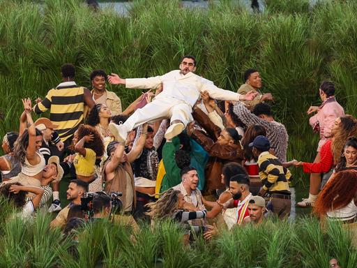 Eine Gruppe von Tänzerinnen und Tänzern steht vor Zuckerrohrpflanzen und trägt Bad Bunny über sich 