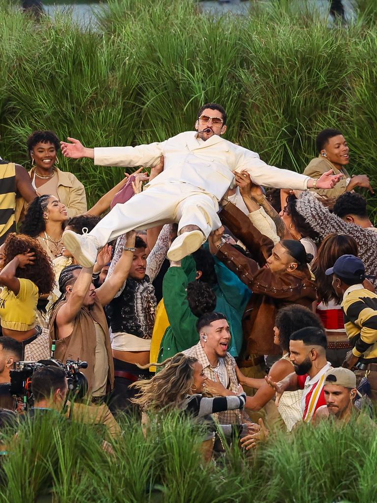 Eine Gruppe von Tänzerinnen und Tänzern steht vor Zuckerrohrpflanzen und trägt Bad Bunny über sich 