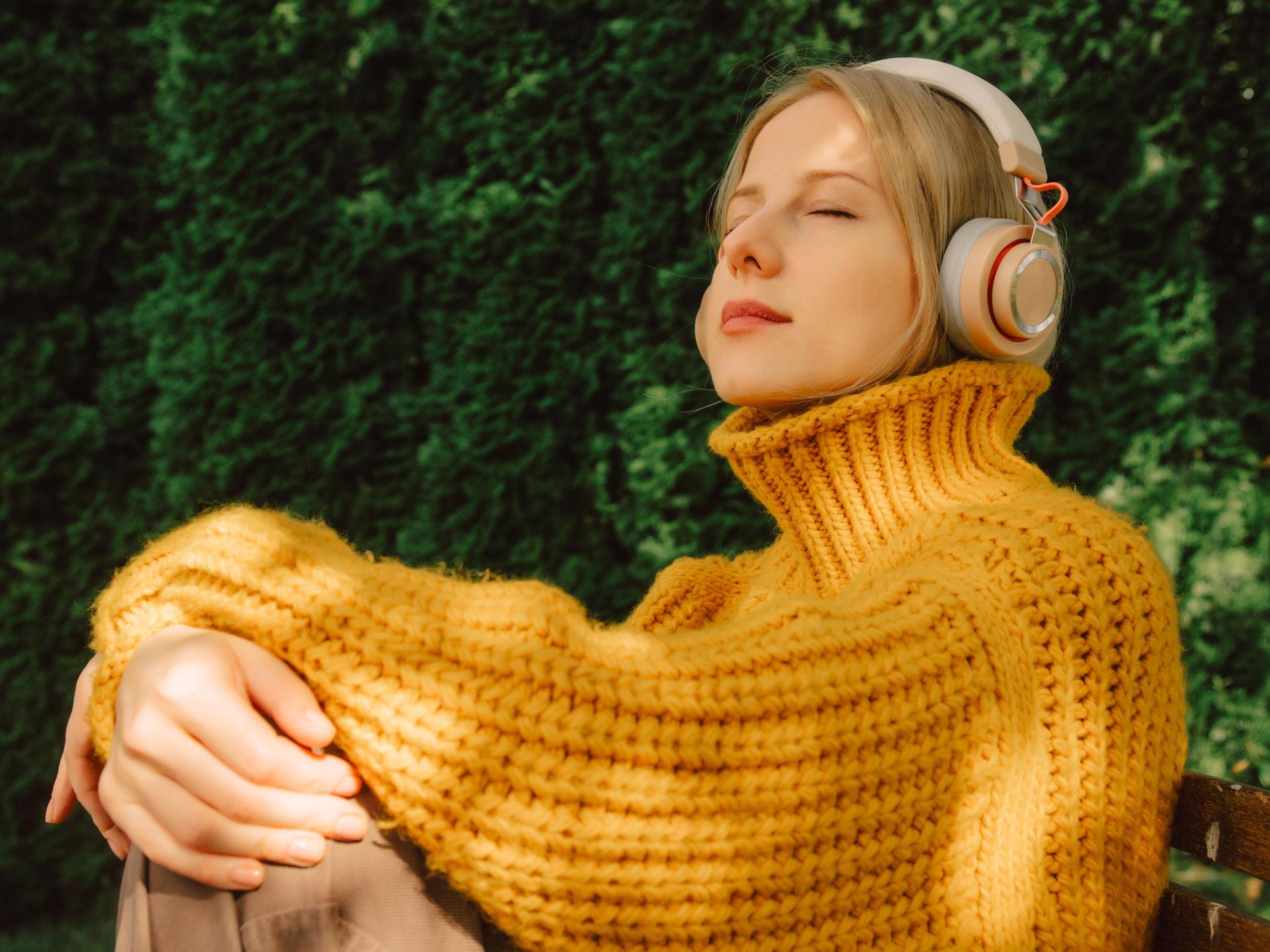 Eine Frau in einem gelben Sweeter sitzt entspannt auf einer Bank im Park und hört genussvoll Musik mit ihren Kopfhörern. Eine Frau in einem gelben Sweeter sitzt entspannt auf einer Bank im Park und hört genussvoll Musik mit ihren Kopfhörern.