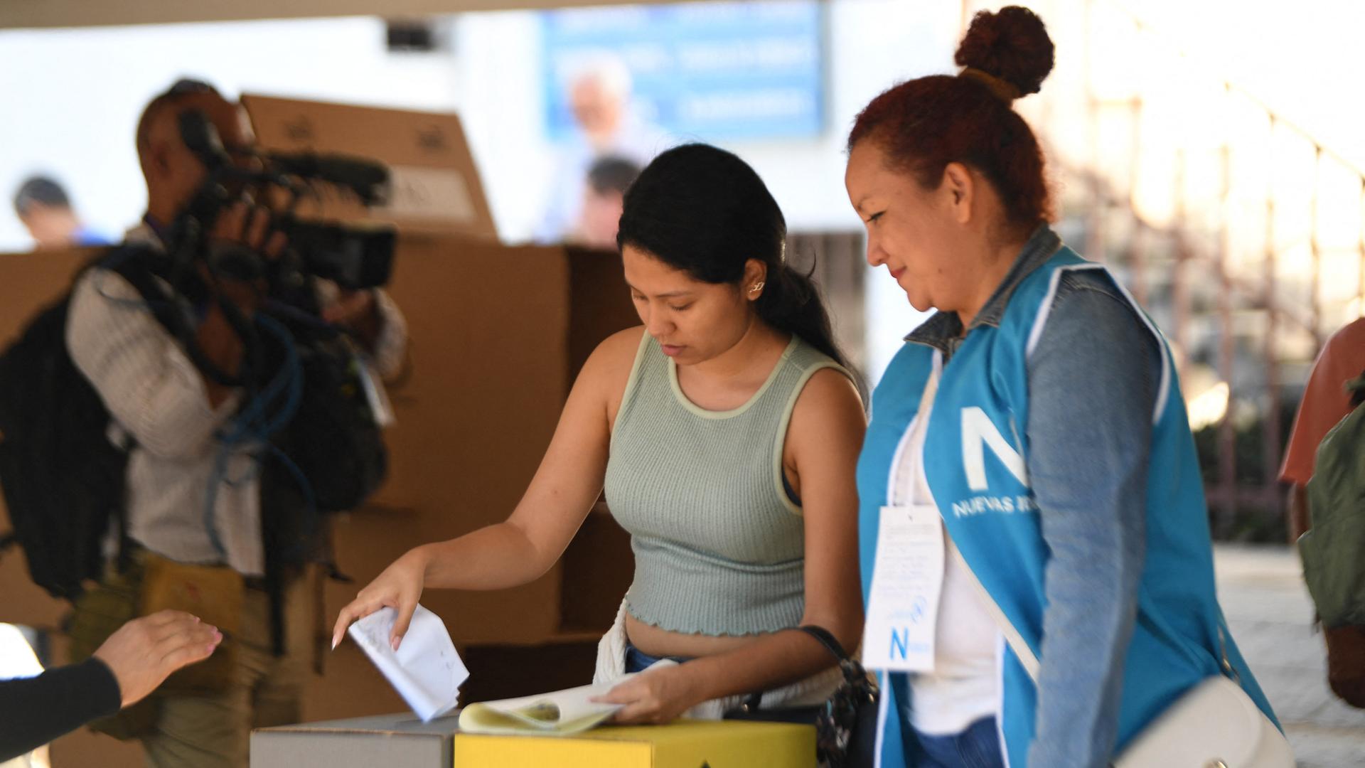 Eine Frau wirft in El Salvador ihren Wahlzettel in die Wahlurne. Eine zweite Frau steht daneben.