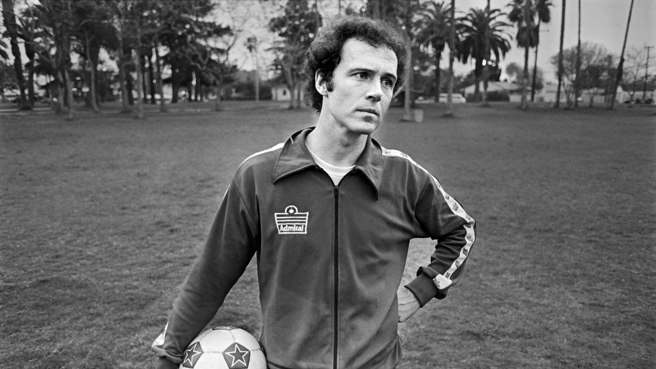 Franz Beckenbauer steht auf einer Wiese vor Palmen in Kalifornien, er hält einen Fußball im rechten Arm.