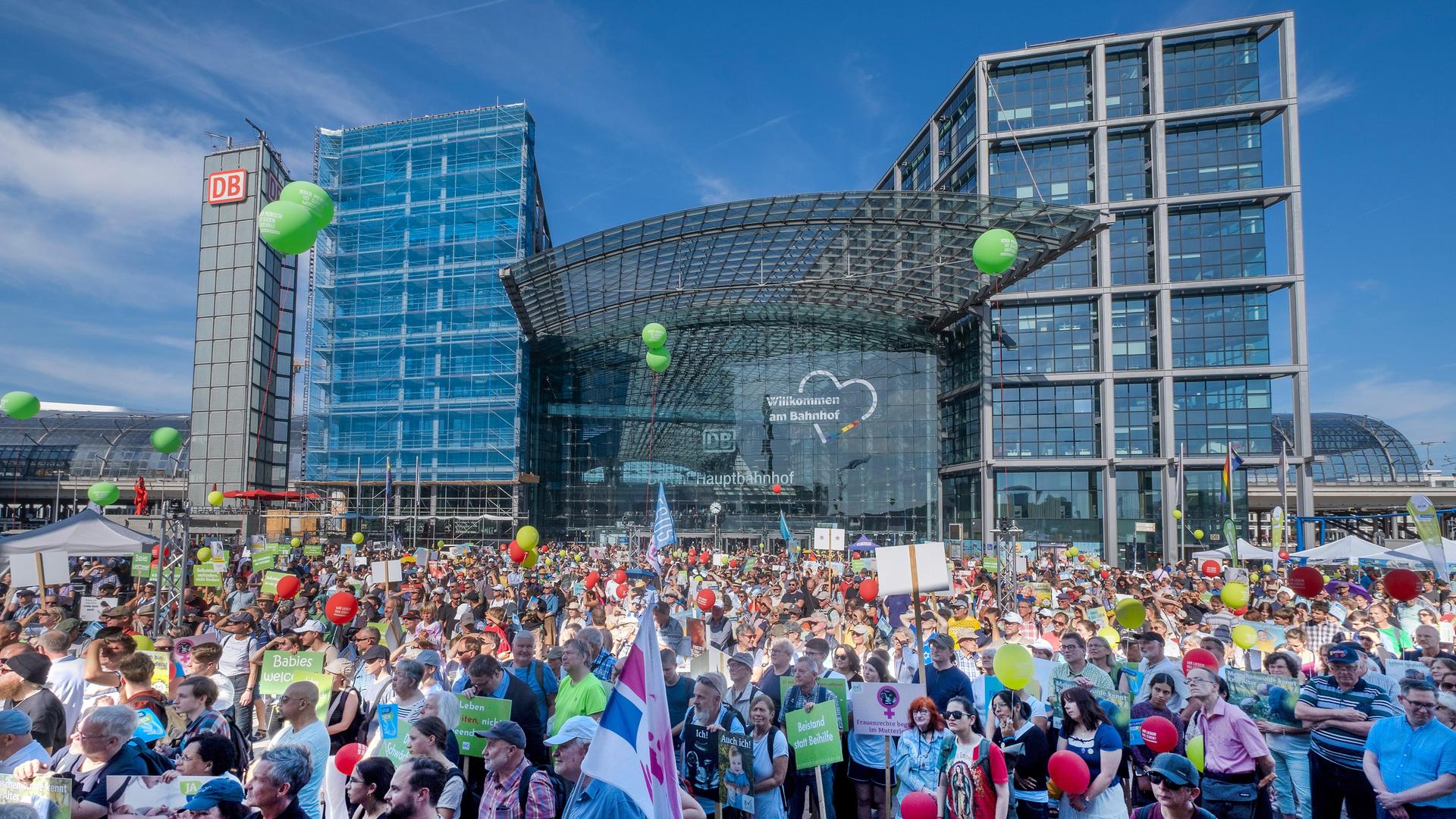 Das Bild zeigt viele Menschen vor dem Berliner Hauptbahnhof bei strahlend blauem Himmel. Über der Menge sind grüne und rote Luftballons zu erkennen, einige halten Plakate oder Fahnen in der Hand. Das Bild zeigt viele Menschen vor dem Berliner Hauptbahnhof bei strahlend blauem Himmel. Über der Menge sind grüne und rote Luftballons zu erkennen, einige halten Plakate oder Fahnen in der Hand.