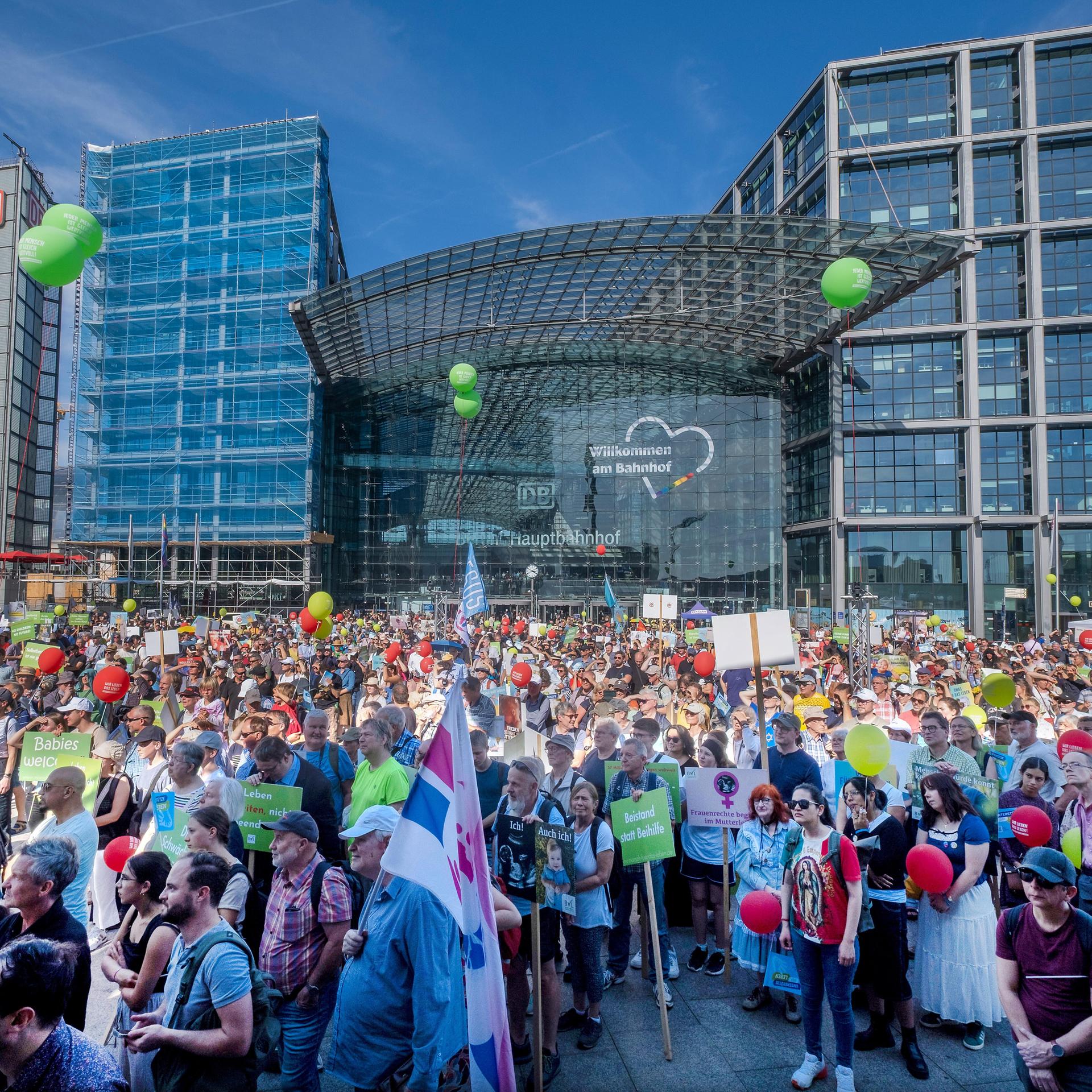 Das Bild zeigt viele Menschen vor dem Berliner Hauptbahnhof bei strahlend blauem Himmel. Über der Menge sind grüne und rote Luftballons zu erkennen, einige halten Plakate oder Fahnen in der Hand.