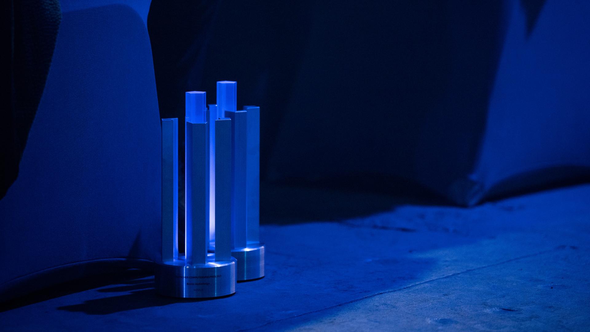 Zwei Preise stehen auf dem Boden - Säulen auf einer Plattform. Es sind die Deutschen Computerspielpreise. Die Szene ist in blaues Licht getaucht.