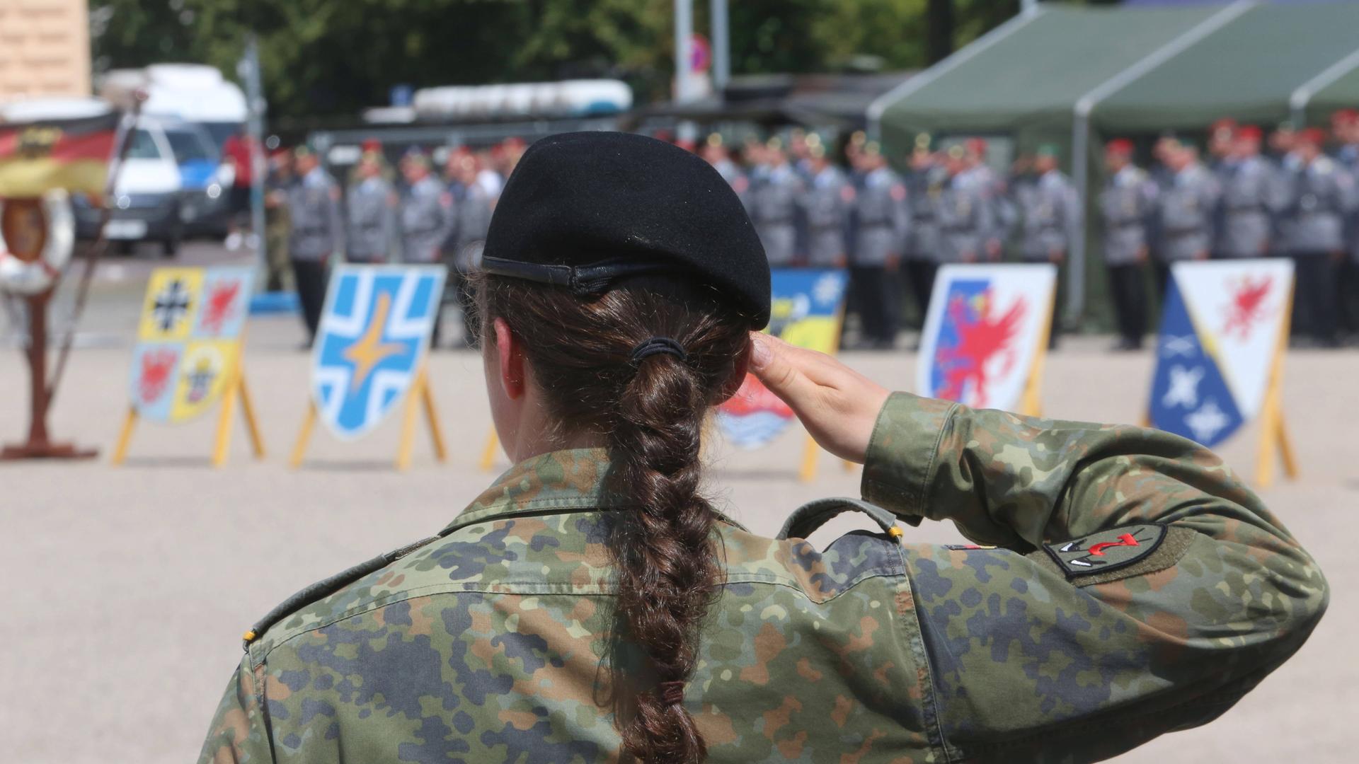 Eine Soldatin der Bundeswehr steht mit dem Rücken zum Bild. Sie hat einen schwarzen langen geflochtenen Zopf und hebt den rechten Arm nach oben und die Hand an die Stirn. Sie trägt eine Uniform. 