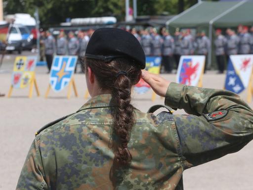 Eine Soldatin der Bundeswehr steht mit dem Rücken zum Bild. Sie hat einen schwarzen langen geflochtenen Zopf und hebt den rechten Arm nach oben und die Hand an die Stirn. Sie trägt eine Uniform. 