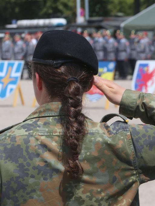 Eine Soldatin der Bundeswehr steht mit dem Rücken zum Bild. Sie hat einen schwarzen langen geflochtenen Zopf und hebt den rechten Arm nach oben und die Hand an die Stirn. Sie trägt eine Uniform. 