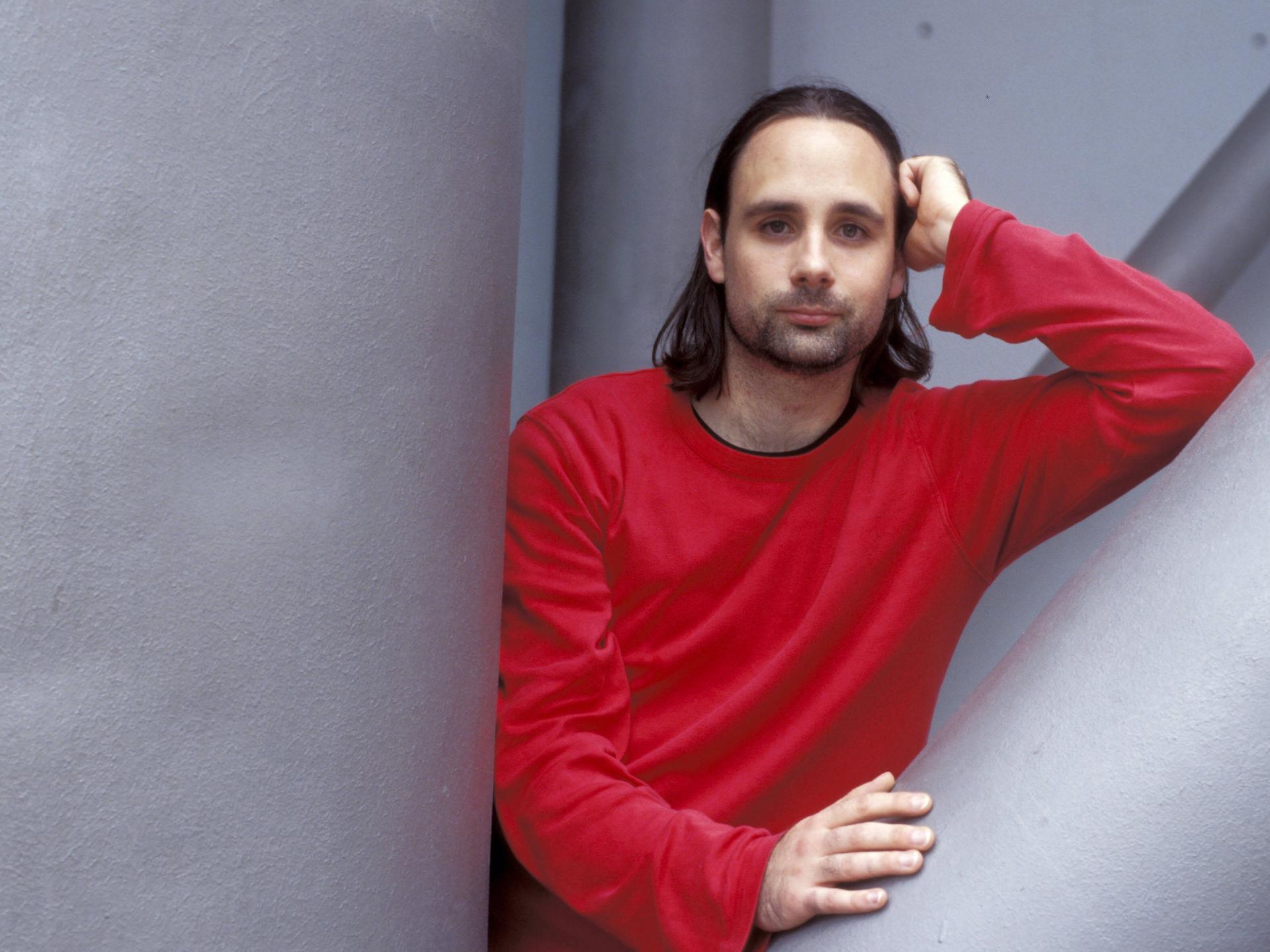 Der Autor Jan Costin Wagner im roten Pullover vor einer grauen Wand in lässiger Pose