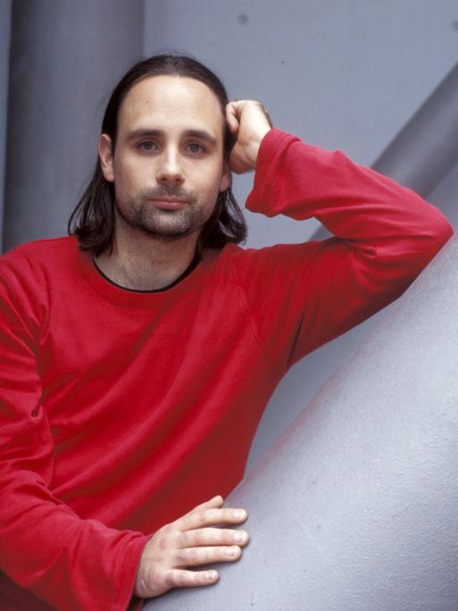 Der Autor Jan Costin Wagner im roten Pullover vor einer grauen Wand in lässiger Pose