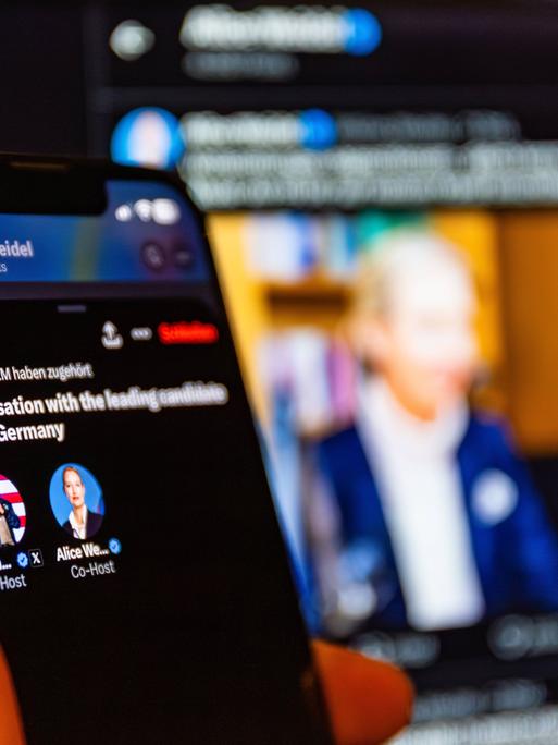 Eine Hand, von der man nur Daumen und Zeigefinger sieht, hält ein Handy mit der Ankündigung von der Unterhaltung von Elon Musk und Alice Weidel mit ihren X-Profilfotos. Im Hintergrund angeschnitten und verschwommen ein Foto von Weidel.