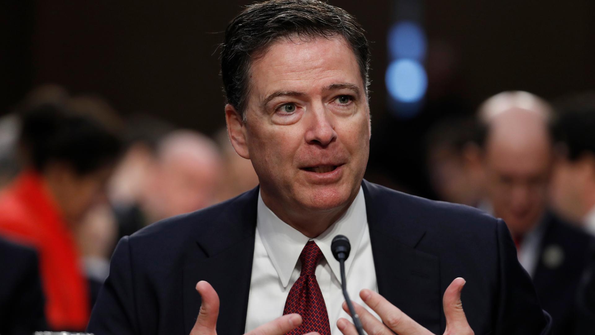 James Comey gestikuliert vor einem Mikrofon mit beiden Händen.