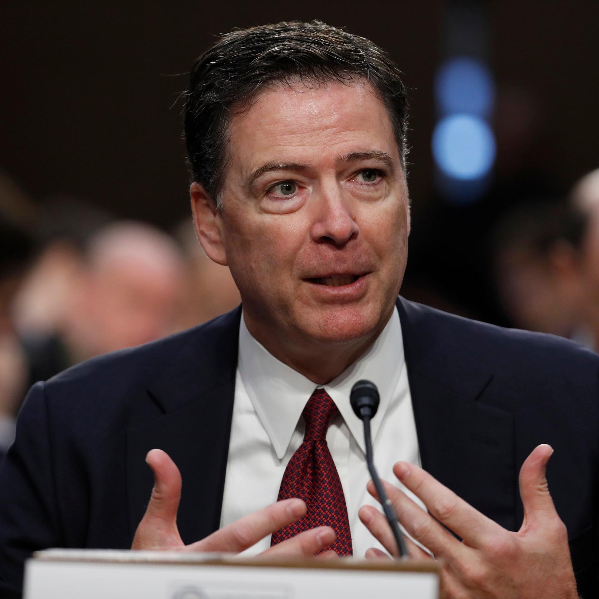 James Comey gestikuliert vor einem Mikrofon mit beiden Händen.