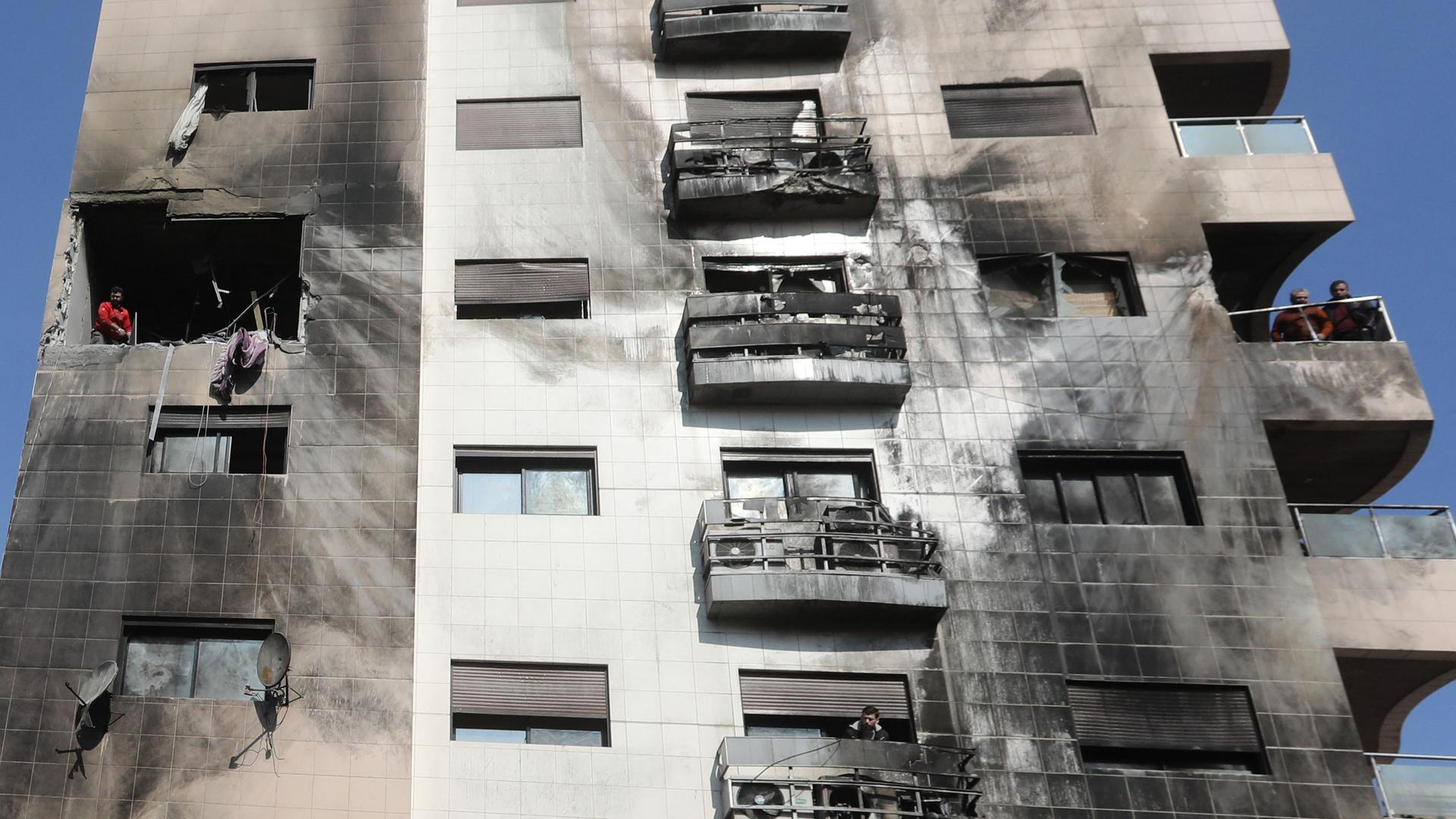 Menschen untersuchen die Schäden an einem Gebäude nach einem israelischen Angriff. Fenster sind zerstört, die Hauswand ist durch Rauch schwarz gefärbt.