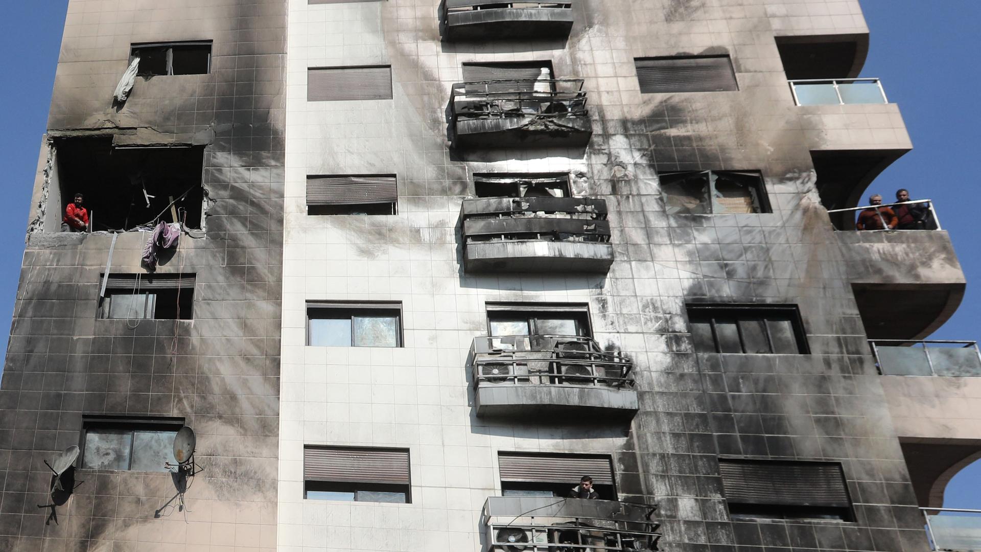 Syrien, Damaskus: Menschen untersuchen die Schäden an einem Gebäude nach einem israelischen Angriff. Fenster sind zerstört, die Hauswand ist durch Rauch schwarz gefärbt. 