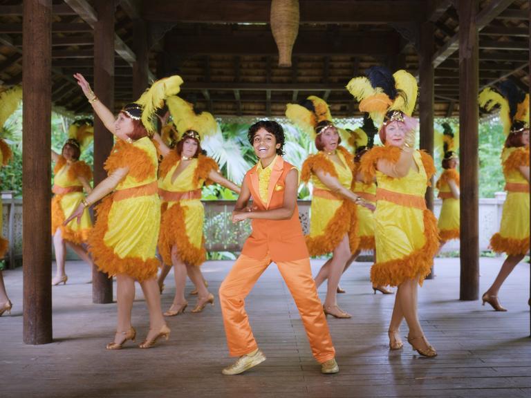 Ein junge in gelb-oranger Kleidung tanzt mit vielen gleichfalls in gelb-orange gekleideten Frauen in einer Pagode.
