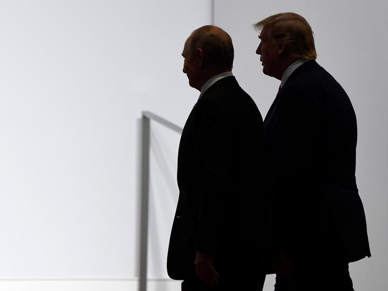 US-Präsident Donald Trump und Russlands Präsident Wladimir Putin stehen dicht beieinander vor einer weißen Wand. Sie sind im Profil und im Halbschatten abgebildet. 