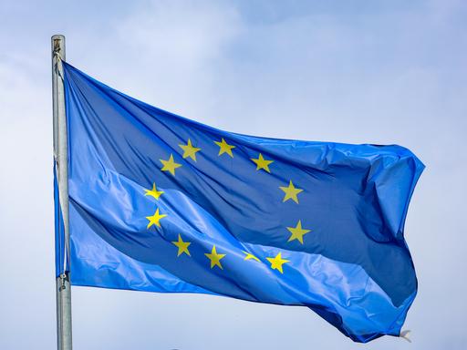 Eine EU-Flagge, die 12 Sterne auf blauem Grund zeigt, weht an einem Fahnenmast im Wind. 