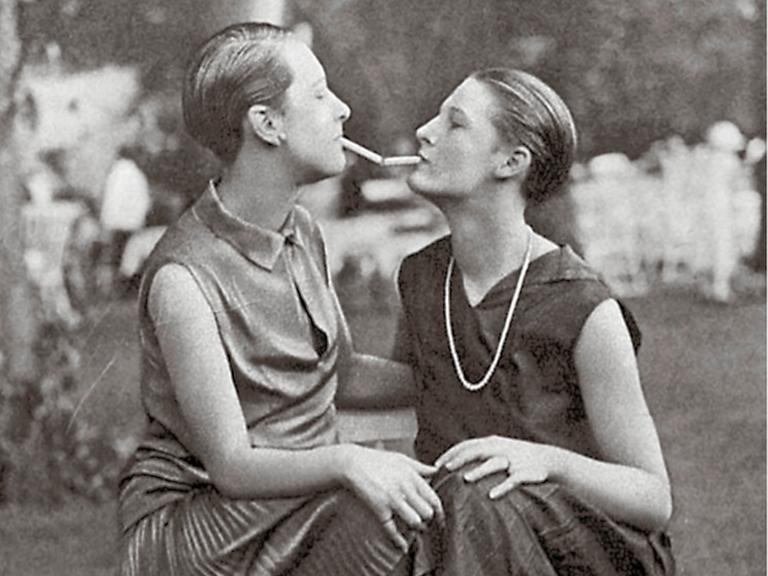Historisches Foto aus den 1920er Jahren von zwei Frauen mit Kurzhaarschnitt. Die eine gibt der Anderen mit ihrer Zigarettenspitze Feuer.