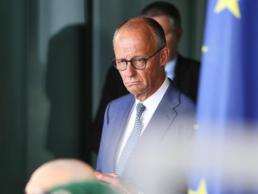 Friedrich Merz im Kanzleramt. er guckt etwas mürrisch. Rechts von ihm ist auf dem Bild etwas unscharf eine Europafahne zu sehen.