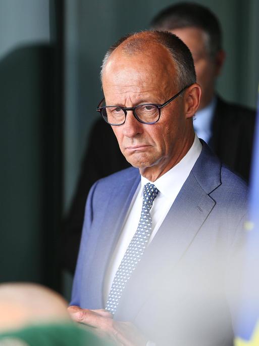Friedrich Merz im Kanzleramt. er guckt etwas mürrisch. Rechts von ihm ist auf dem Bild etwas unscharf eine Europafahne zu sehen.