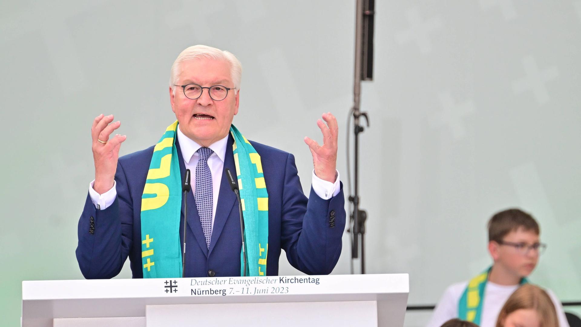 Bundespräsident Steinmeier steht beim Deutschen Evangelischen Kirchentag in Nürnberg hinter einem Rednerpult und hebt beide Hände. Er trägt einen Schal mit dem Motto des Kirchentages, "Jetzt ist Zeit".
