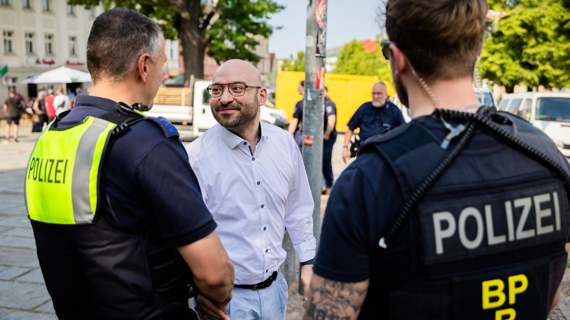 Der Minister im weißen Hemd mit Glatze und Bart sowie Brille steht lächelnd zwischen zwei Polizisten auf dem Platz. Dahinter weitere Personen. Der Minister im weißen Hemd mit Glatze und Bart sowie Brille steht lächelnd zwischen zwei Polizisten auf dem Platz. Dahinter weitere Personen.