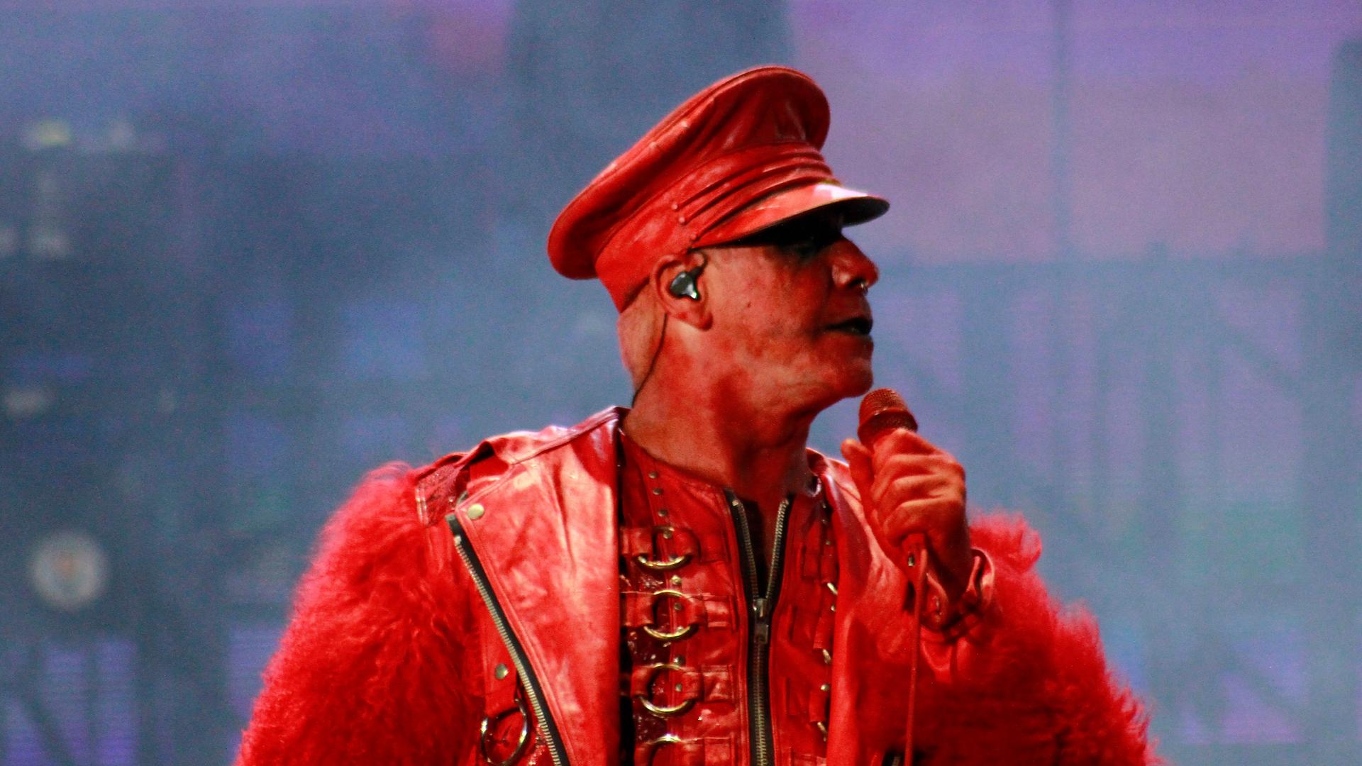 Till Lindemann ist komplett in rot gekleidet, auch sein Gesicht ist rot geschminkt. Er hält ein Mikrofon in der Hand und steht dabei auf einer Bühne.