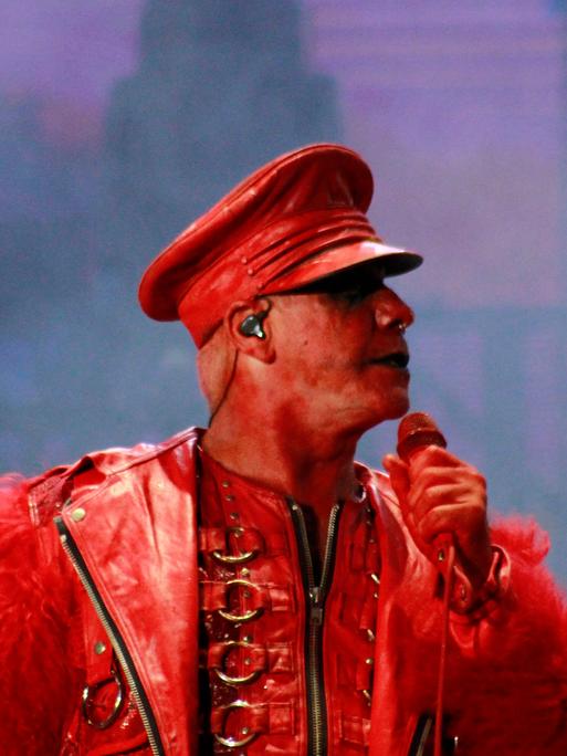 Till Lindemann ist komplett in rot gekleidet, auch sein Gesicht ist rot geschminkt. Er hält ein Mikrofon in der Hand und steht dabei auf einer Bühne.