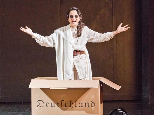 Im Bild: Schauspielerin Sesede Terziyan im Stück "Hätte klappen können - Ein patriotischer Liederabend" von Ruud Gielens im Maxim Gorki Theater in Berlin. Sie springt aus einem Pappkarton auf dem in Frakturschrift "Deutschland" steht.