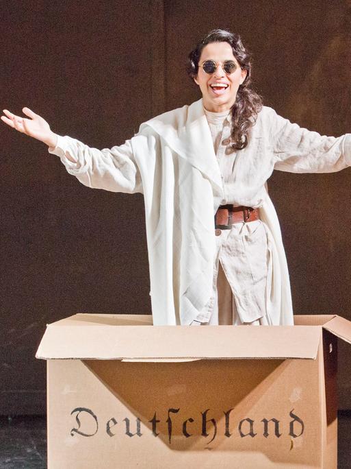 Im Bild: Schauspielerin Sesede Terziyan im Stück "Hätte klappen können - Ein patriotischer Liederabend" von Ruud Gielens im Maxim Gorki Theater in Berlin. Sie springt aus einem Pappkarton auf dem in Frakturschrift "Deutschland" steht.