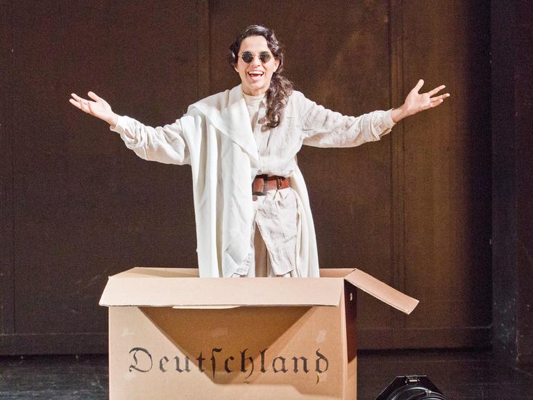 Im Bild: Schauspielerin Sesede Terziyan im Stück "Hätte klappen können - Ein patriotischer Liederabend" von Ruud Gielens im Maxim Gorki Theater in Berlin. Sie springt aus einem Pappkarton auf dem in Frakturschrift "Deutschland" steht.