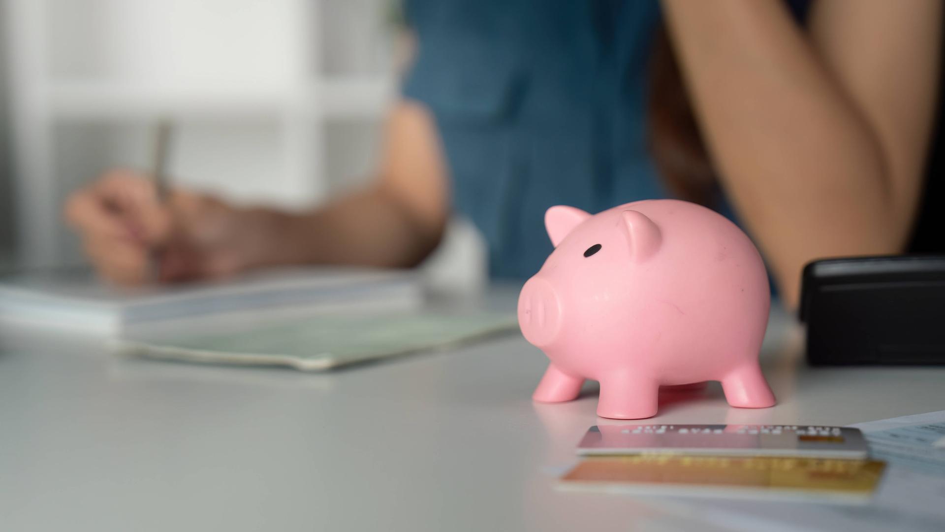 Eine Frau im Hintergrund sitzt am Schreibtisch und schreibt in ein Heft. Im Vordergrund steht ein Sparschwein, Taschenrechner und liegen Kreditkarten.