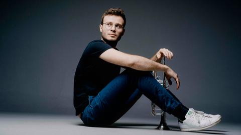 Ein junger Mann sitzt auf dem Boden, zwischen den Beinen steht seine Trompete, dessen Mundstück er lässig mit einer Hand abdeckt. Er trägt ein schwarzes T-Shirt, Jeans und weiße Sneaker.