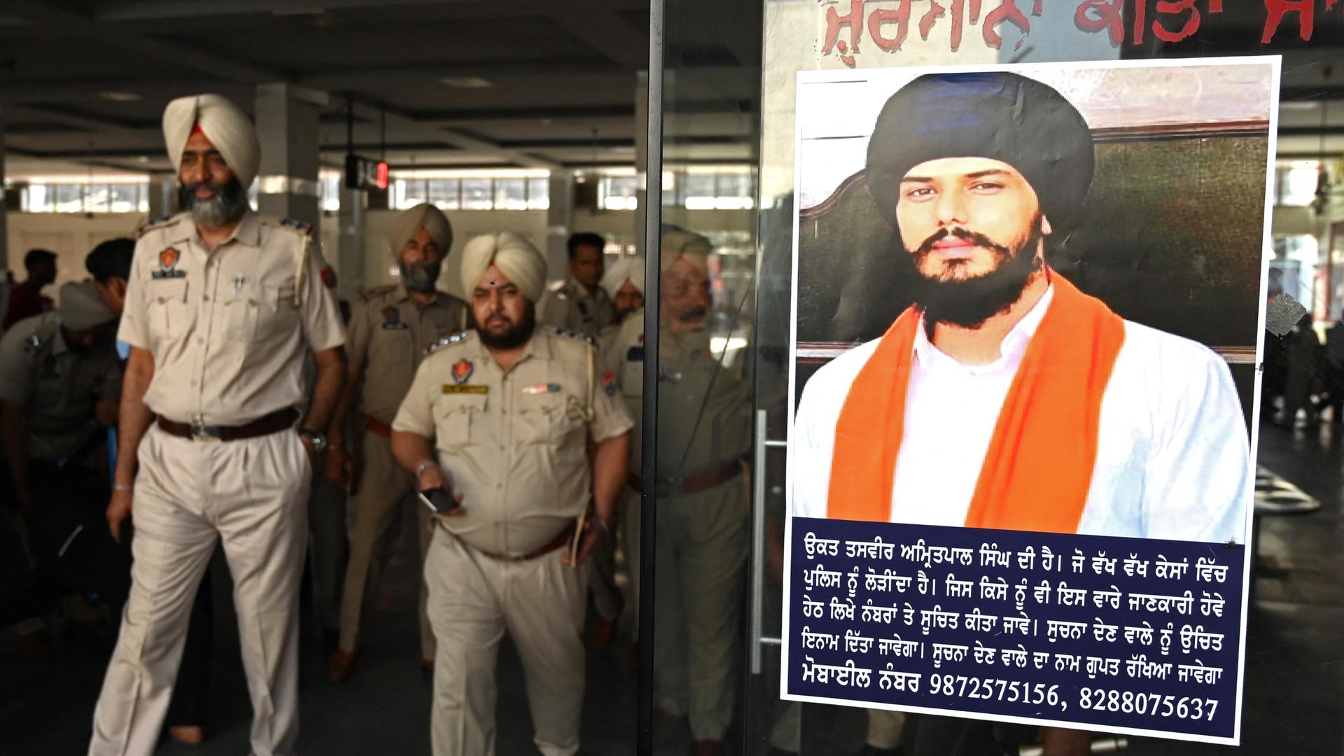 An einem Bahnhof hängt ein Fahndungsplakat mit dem Bild des radikalen Sikh-Predigers Amritpal Singh. Es gehen mehrere Polizisten daran vorbei. An einem Bahnhof hängt ein Fahndungsplakat mit dem Bild des radikalen Sikh-Predigers Amritpal Singh. Es gehen mehrere Polizisten daran vorbei.