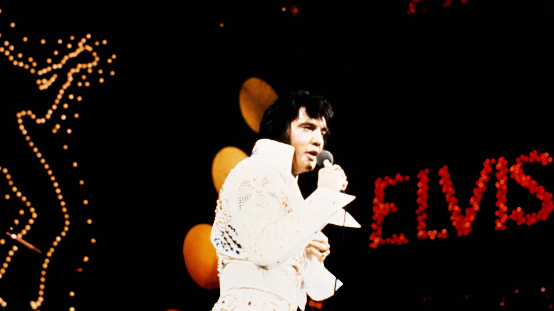 Elvis Presley bei einem Konzert, im Hintergrund auf schwarzem Wand das Wort "ELVIS"