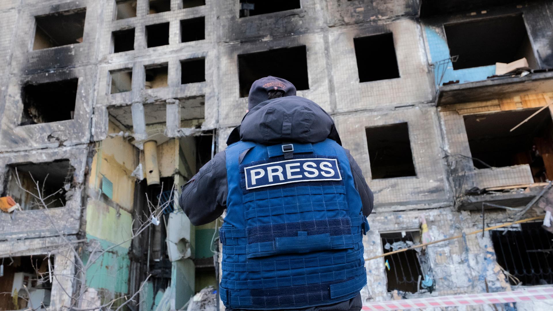 Das Bild zeigt einen Pressevertreter mit einer blauen Weste auf der "Press" in Englisch steht vor einem zerbombten Haus in Kyiv in der Ukraine im Jahr 2022. Das Bild zeigt einen Pressevertreter mit einer blauen Weste auf der "Press" in Englisch steht vor einem zerbombten Haus in Kyiv in der Ukraine im Jahr 2022.