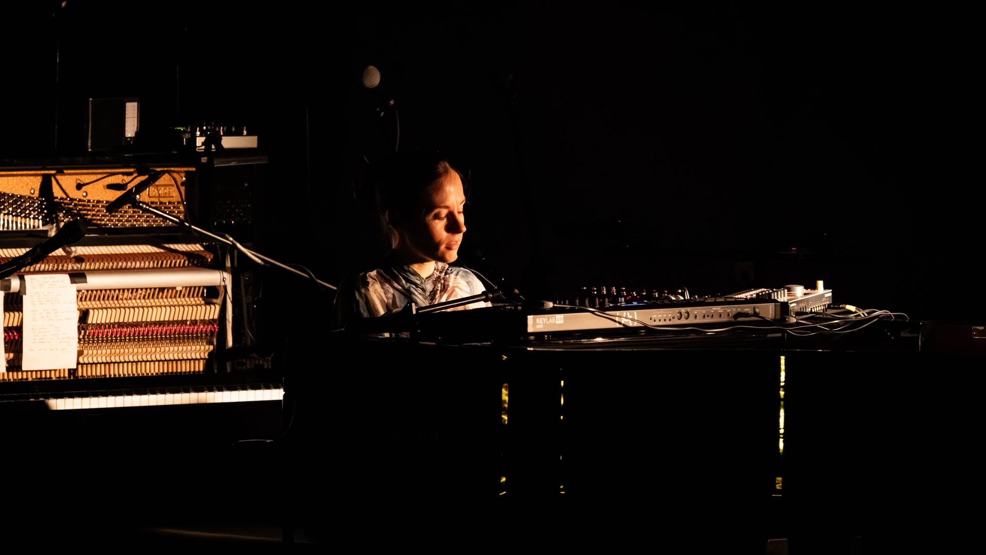 Eine junge Frau sitzt an einem Flügel auf einer Bühne. Der Hintergrund ist überwiegend schwarz, sie sitzt im Licht und wirkt versunken in die Musik. Eine junge Frau sitzt an einem Flügel auf einer Bühne. Der Hintergrund ist überwiegend schwarz, sie sitzt im Licht und wirkt versunken in die Musik.